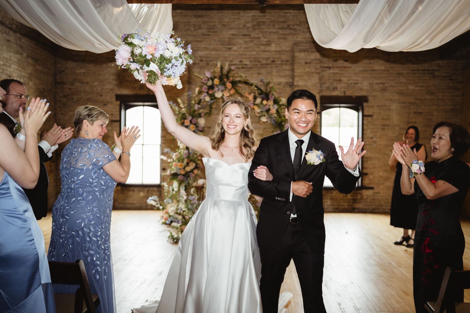 Wedding party celebrating and dancing together on rustic wooden floor with white drapery and floral decor.
