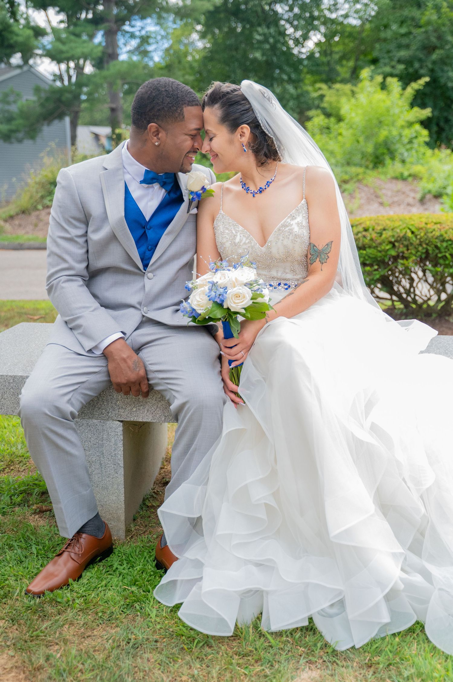 Newlywed couple in gray suit and white gown share tender moment on stone bench in garden setting with blue bouquet.