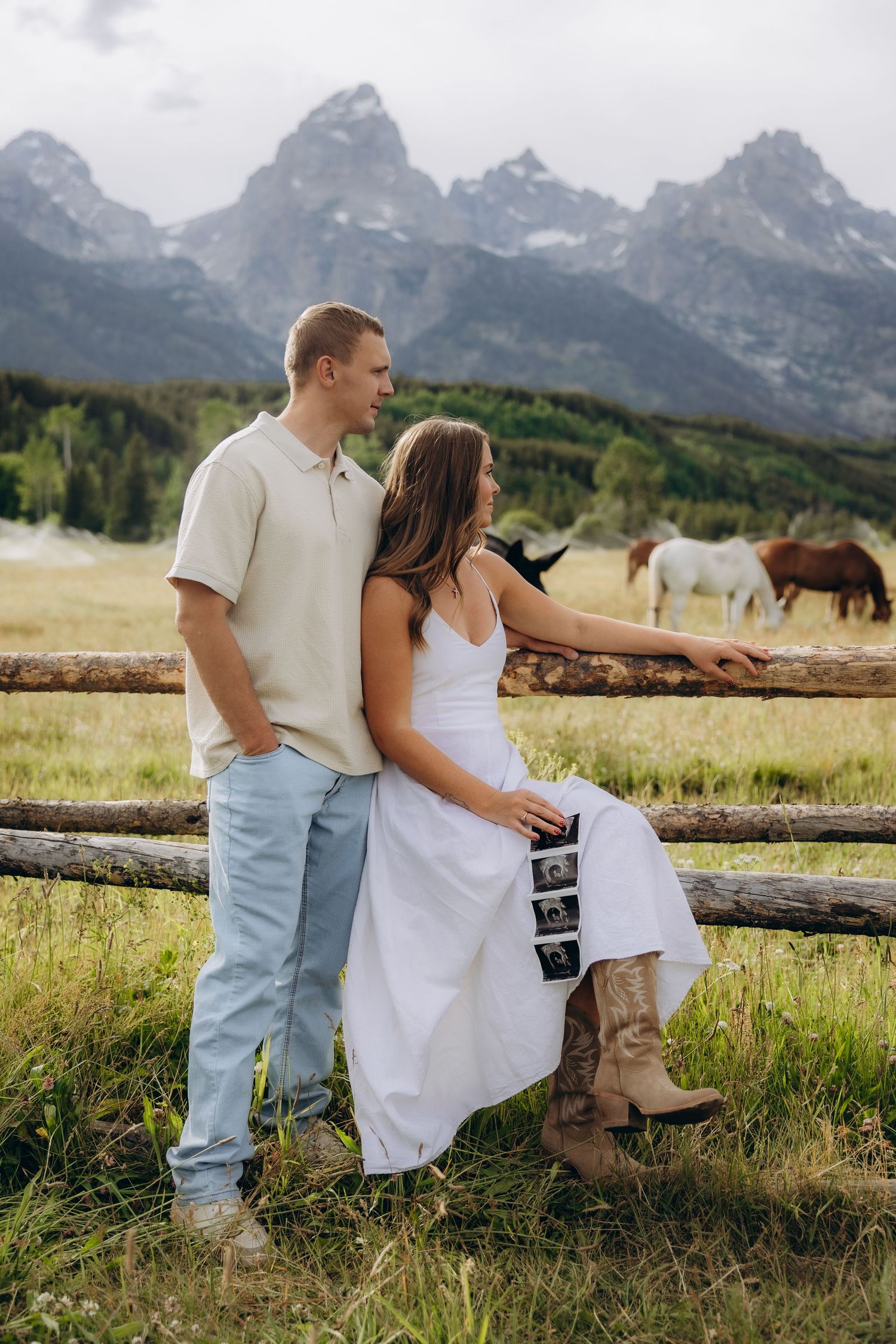 Couple in casual attire sharing an intimate moment on a wooden fence with majestic Teton mountains in the background.