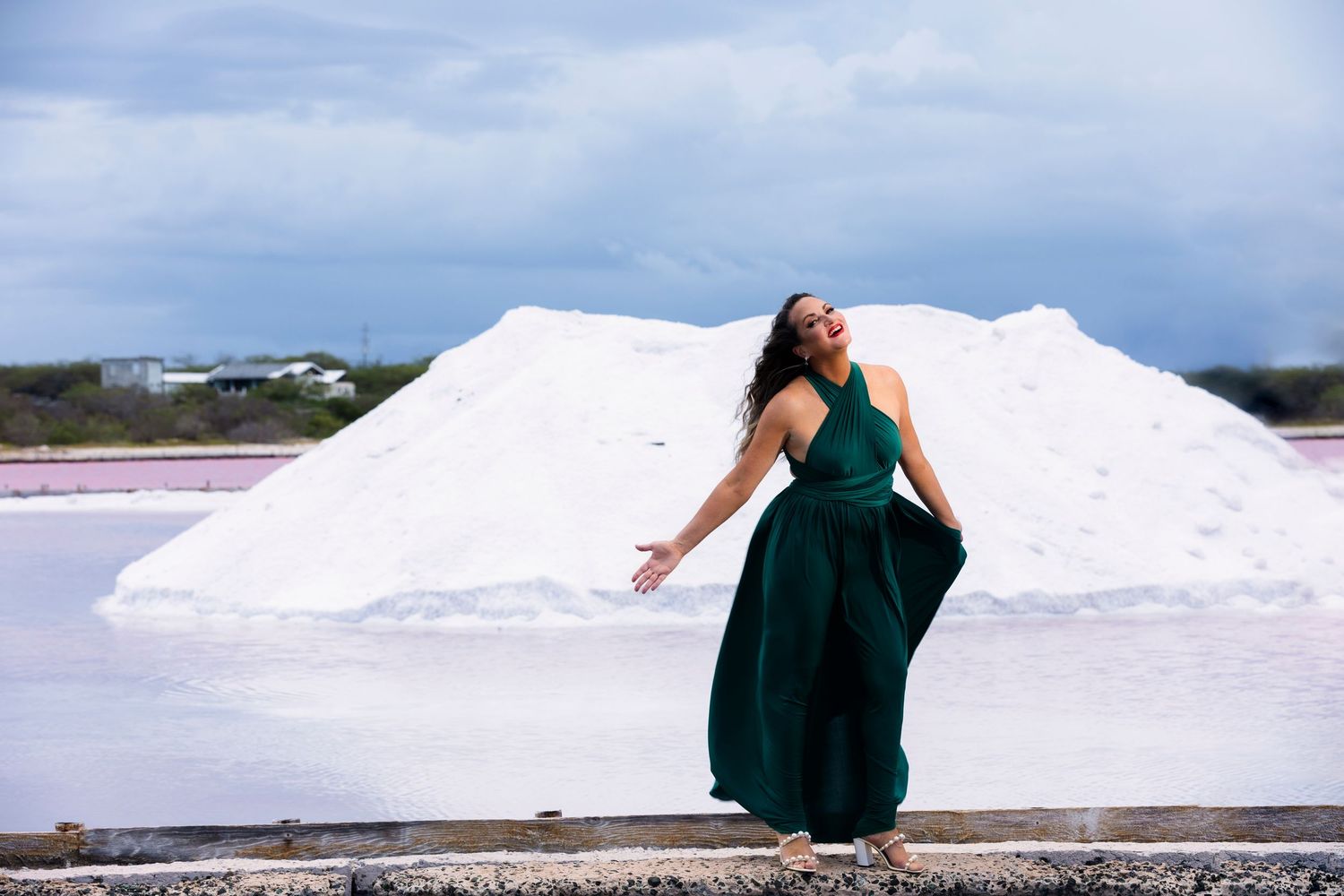 A figure in an emerald green dress poses dramatically against white salt mounds at a salt flat under cloudy skies.