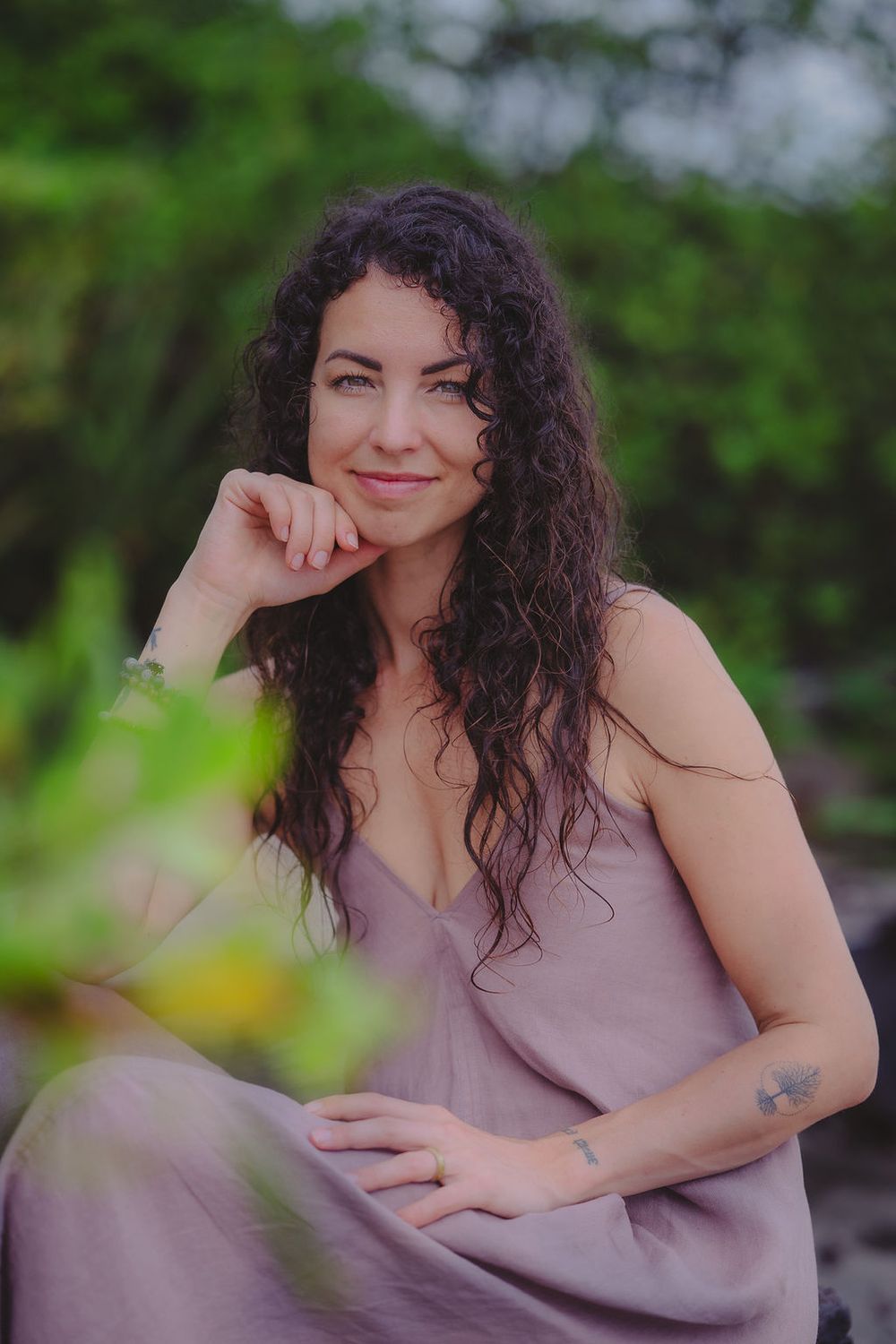 Person in a mauve dress with flowing curly hair poses thoughtfully in an outdoor garden setting.