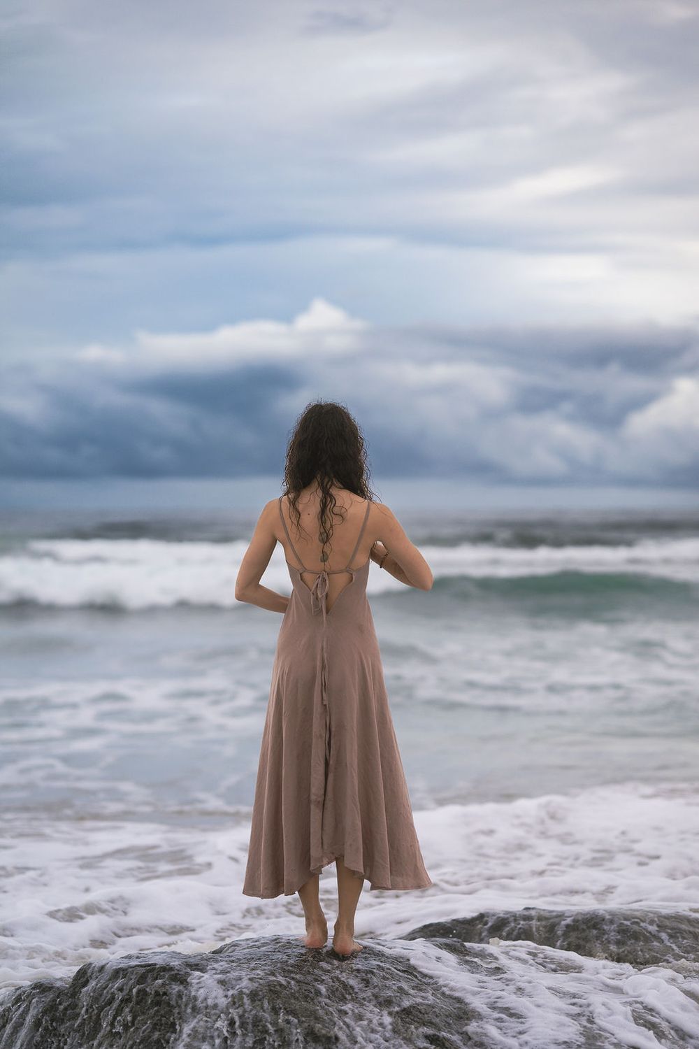 Woman in flowy beige dress stands barefoot on rocky ocean shore under dramatic cloudy sky at dusk.