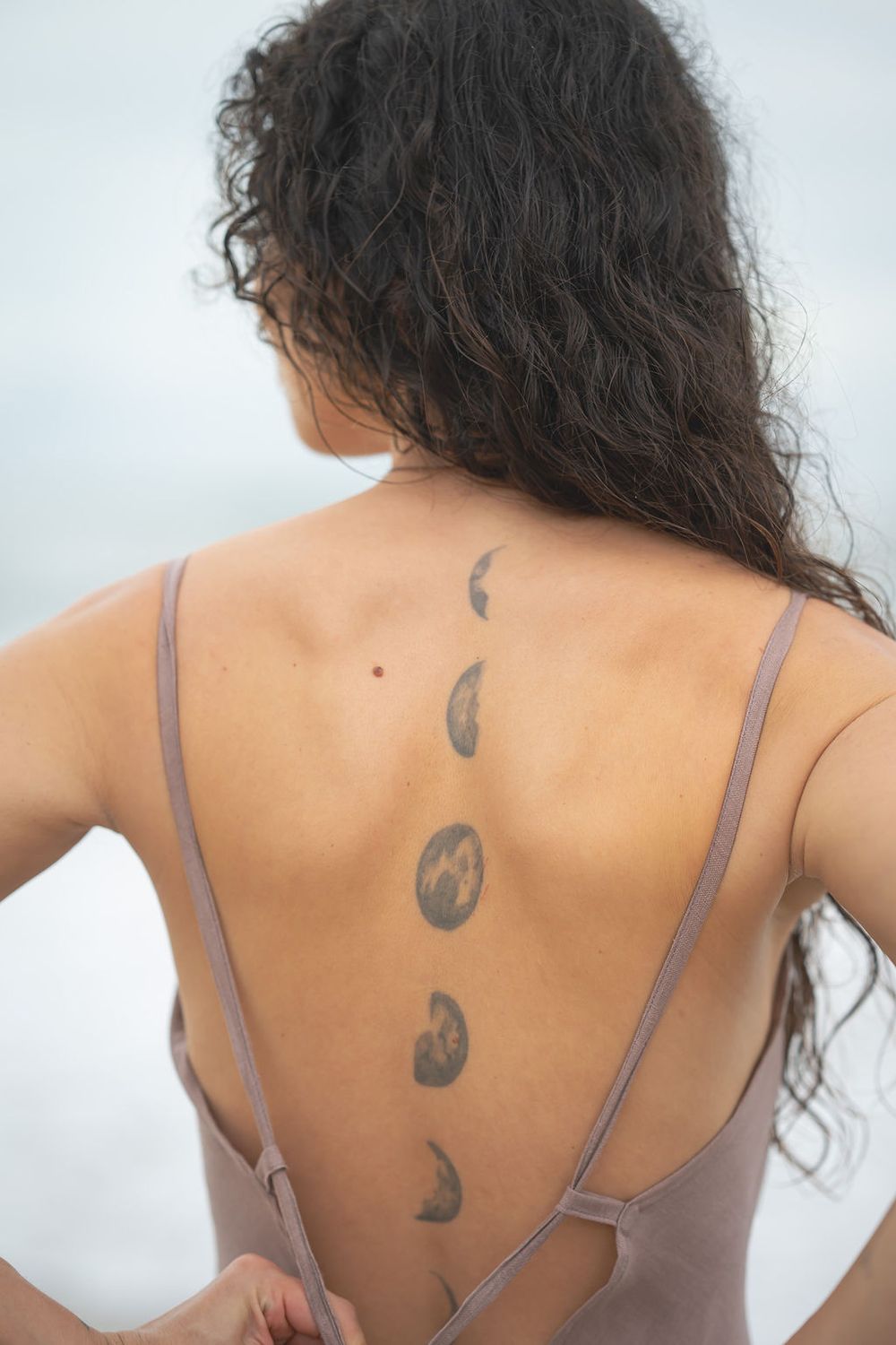 Back view of black curly hair and decorative tattoos seen on a person's shoulder blades and upper back in natural light.