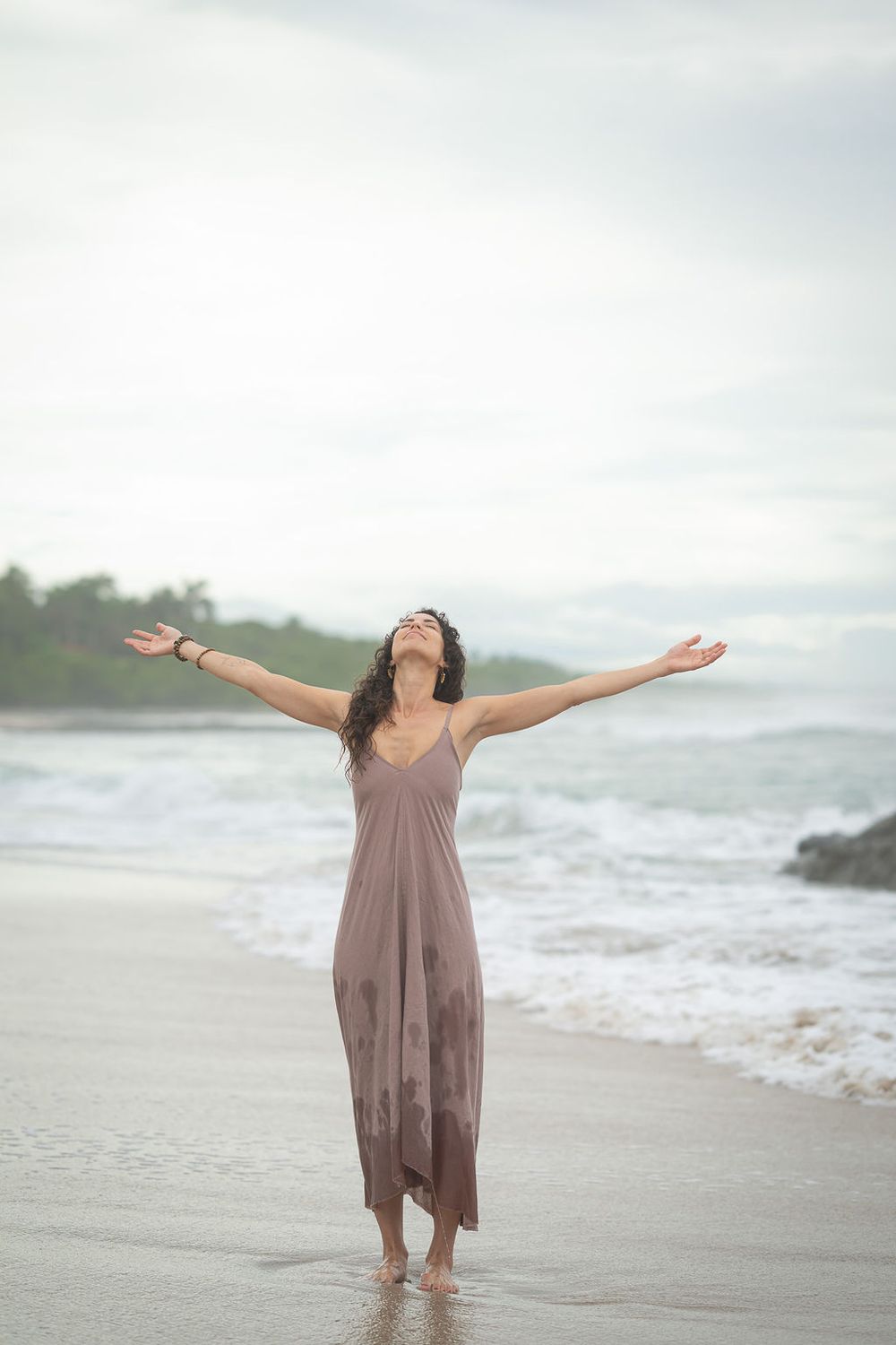 A person in a flowing mauve dress stands with arms outstretched on a misty beach beside crashing waves at the shoreline.