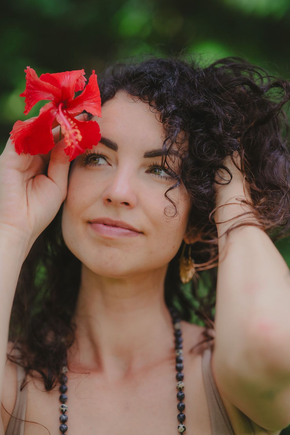 A vibrant red hibiscus flower adorns curly dark hair against a soft green nature backdrop.