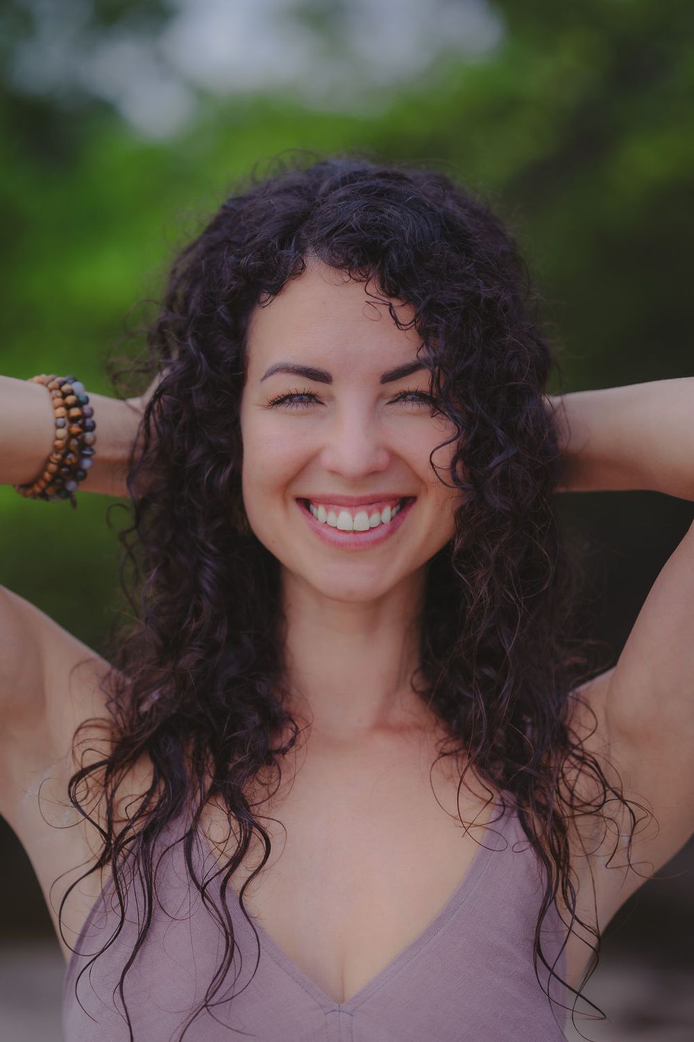 Portrait of a joyful woman with natural curly dark hair wearing a purple top against a blurred green background.