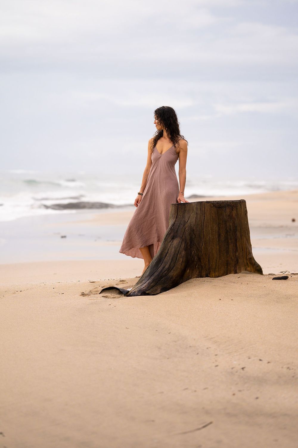 A person wearing a flowing mauve dress sits contemplatively on a large driftwood log at the beach during sunset.