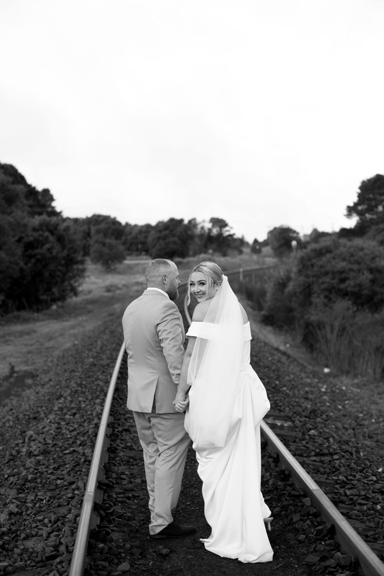 A couple in formal wedding attire embrace on a road with a hillside and trees in the background at sunset.