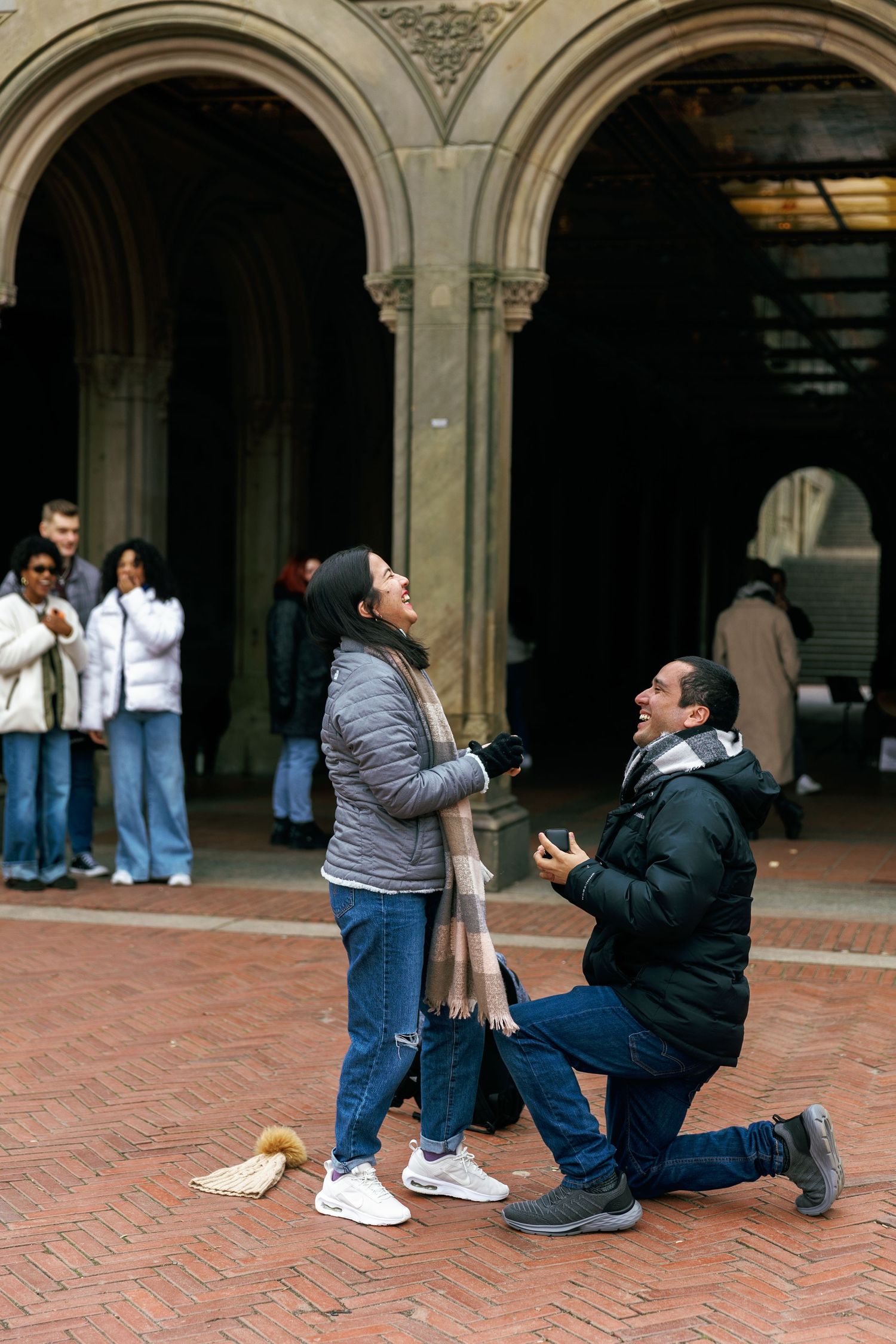 A romantic proposal moment taking place under stone archways in a historic courtyard setting with onlookers in the background.