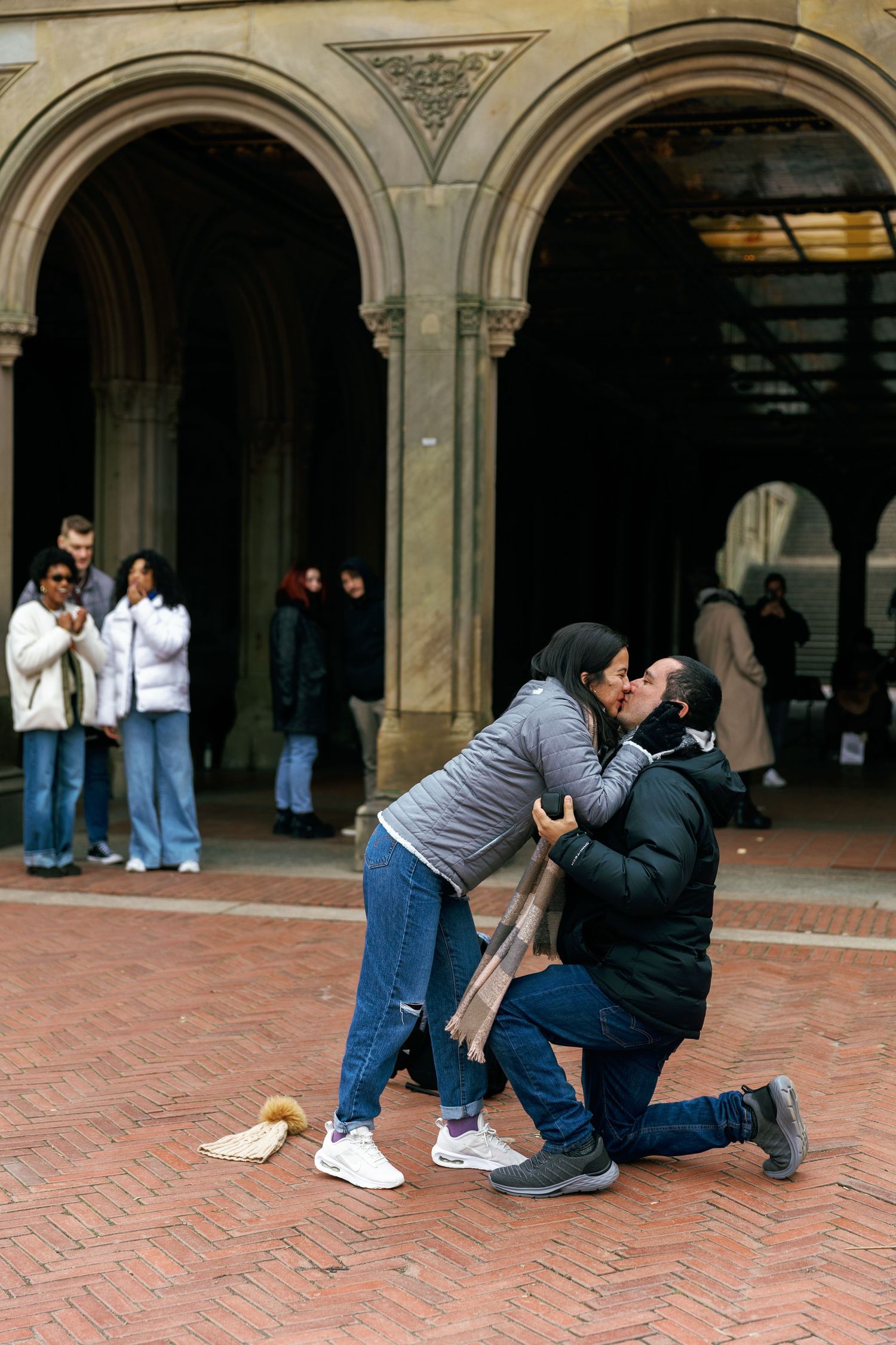 A sequence of four photos showing a proposal taking place under ornate stone archways on a brick courtyard.
