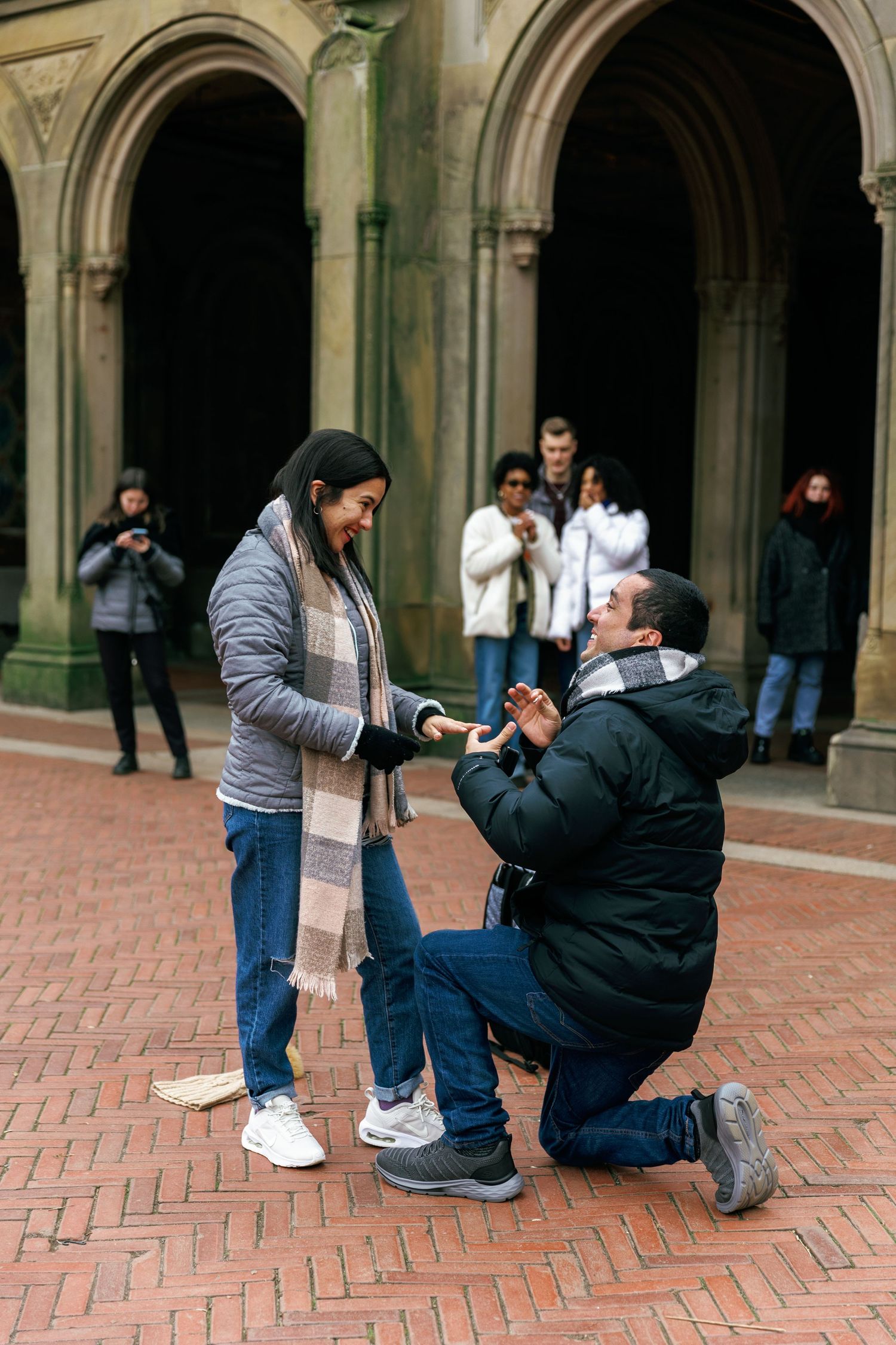 A sequence of photos shows two people during a surprise proposal on a brick courtyard with arched columns in the background.