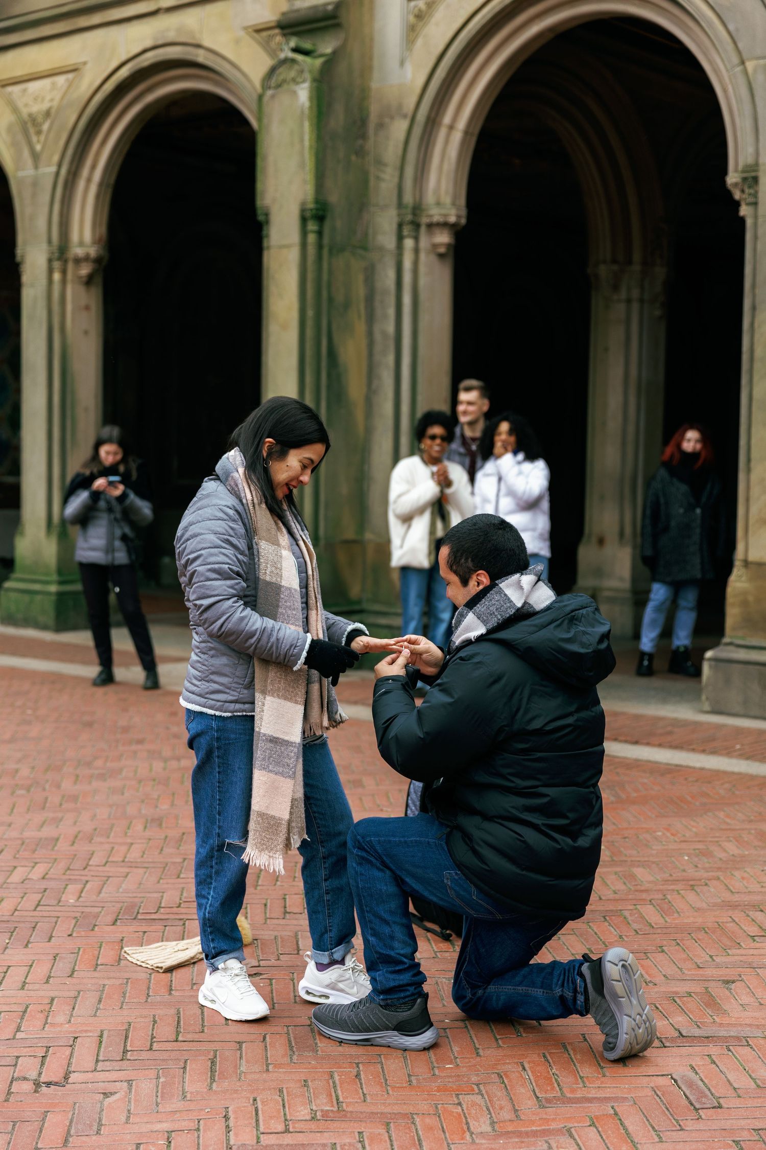 A proposal moment captured in an archway courtyard with someone kneeling on red brick pavement.