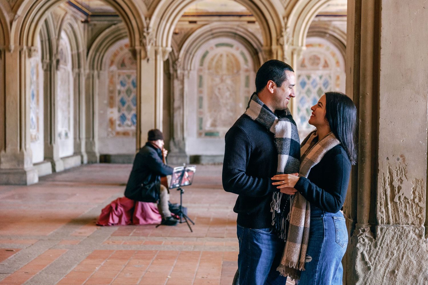 Two people share a close moment under ornate stone archways in a historic corridor setting.