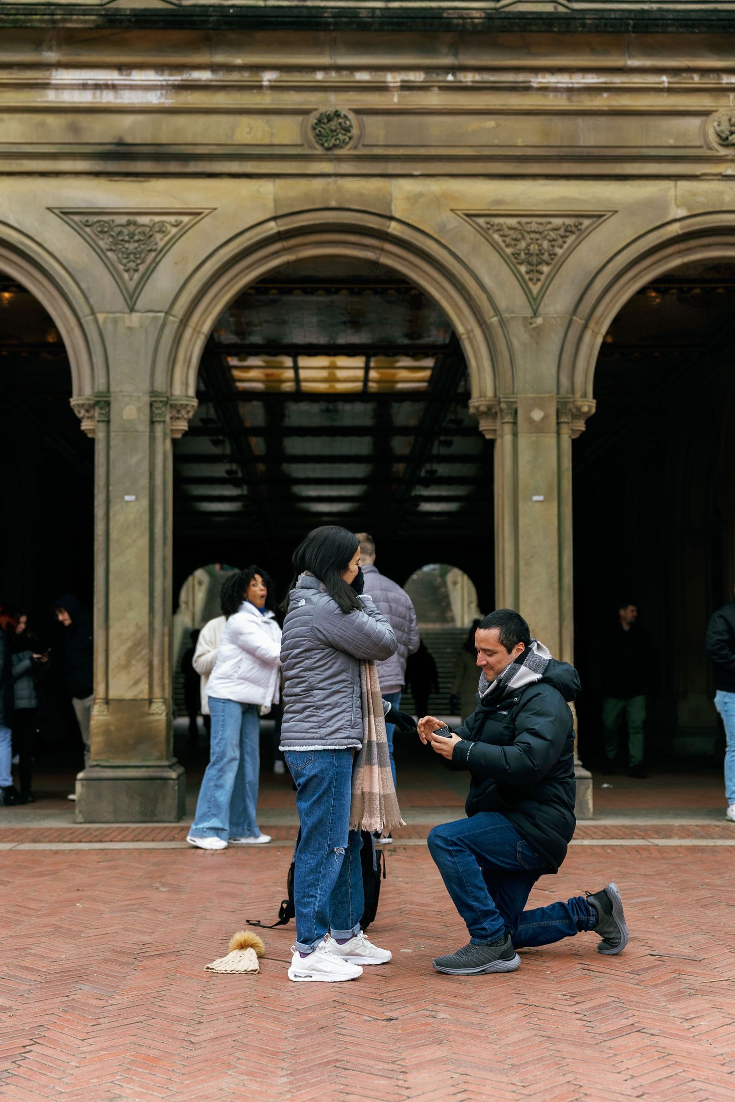 A proposal scene taking place under ornate stone arches at Bethesda Terrace in Central Park, New York City.