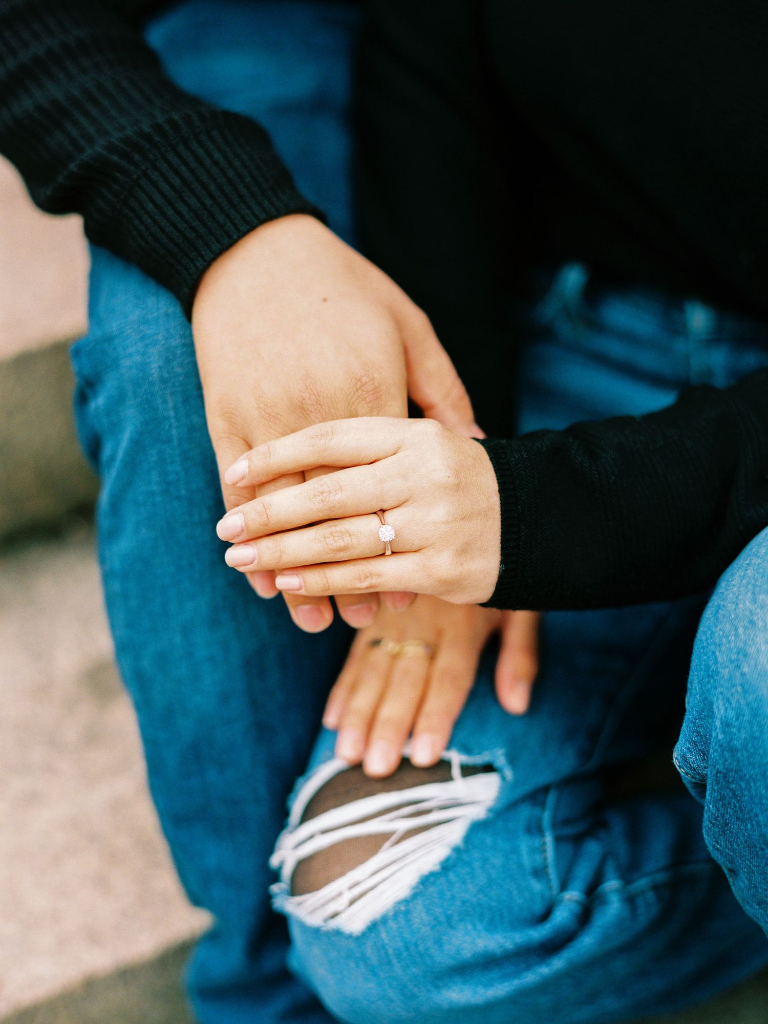 Two people holding hands while sitting together wearing jeans and dark sweaters in intimate moment.