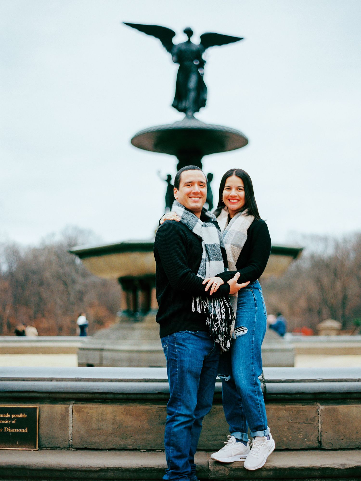 A couple shares a romantic moment while sitting on a bench near a decorative bell and iron railing outdoors.