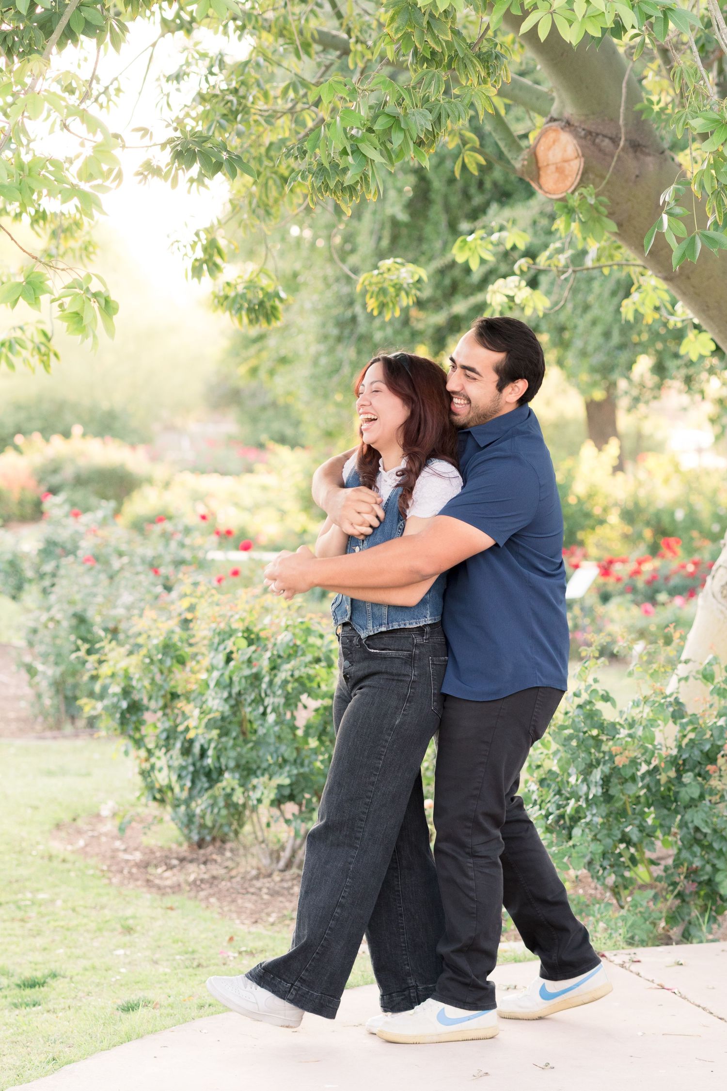 Light and airy portrait of a young couple with the husband hugging his wife from behind under a tall tree in a Mesa, AZ Rose Garden.