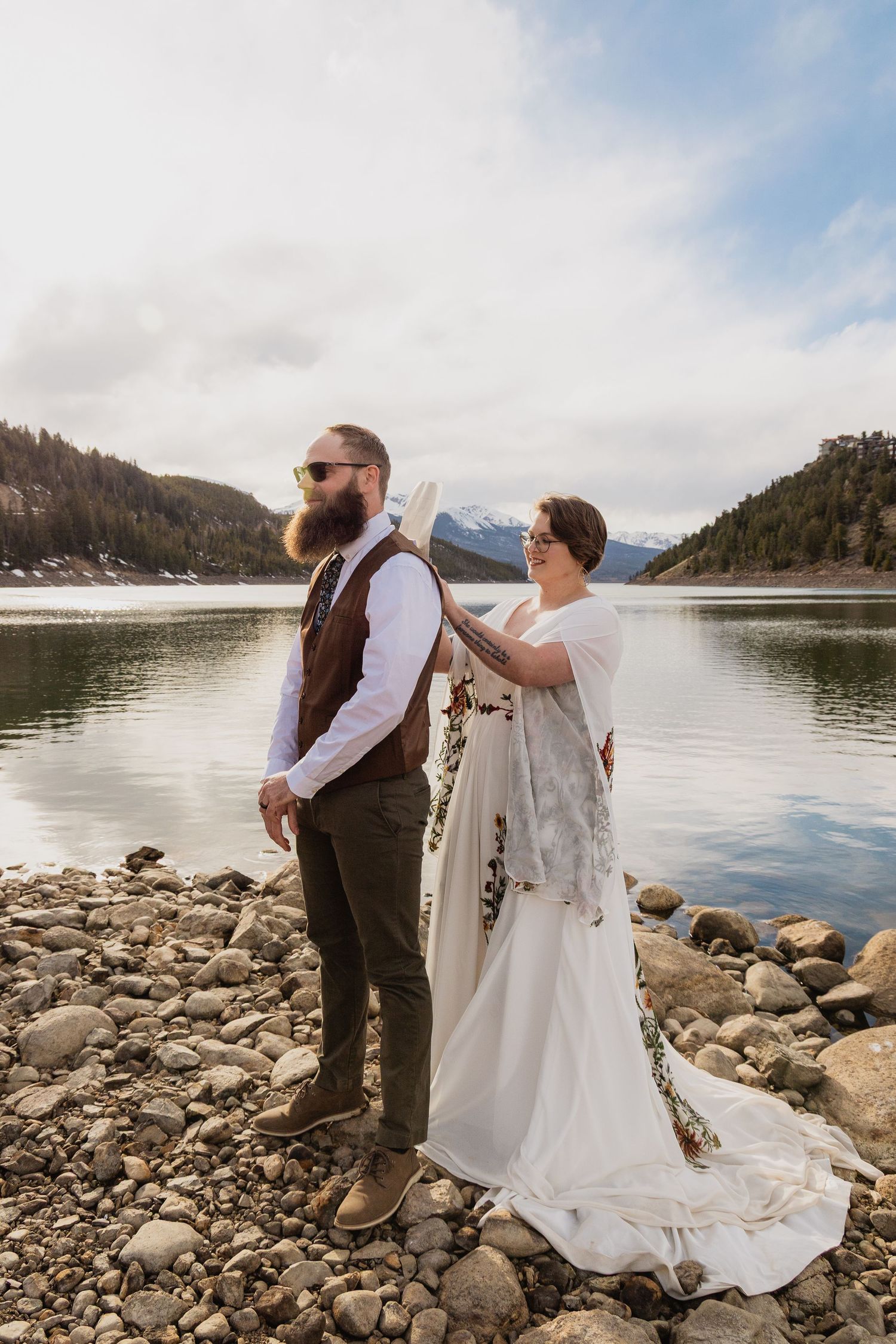 A couple stands on a rocky lakeshore during their mountain wedding with scenic mountains and water in the background.