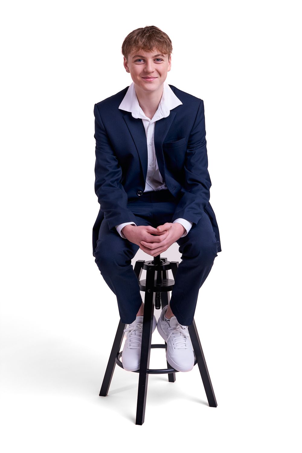 A young person in a navy suit and white dress shirt sits relaxed on a black bar stool against a white background.