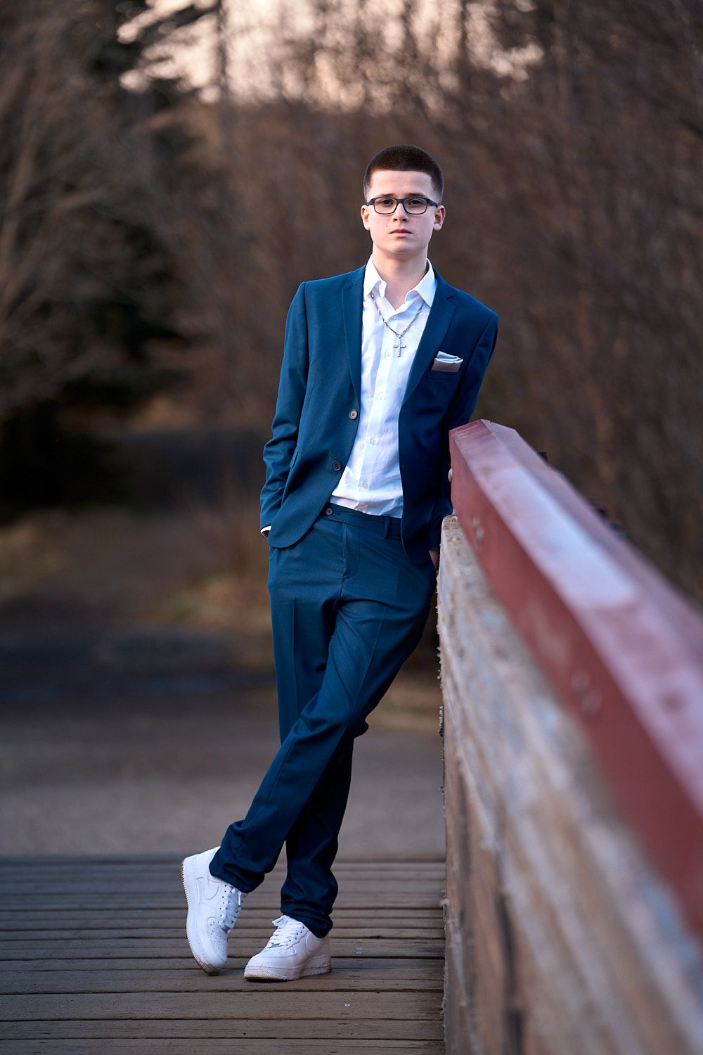 A person in a navy suit and white sneakers poses casually on a wooden bridge.