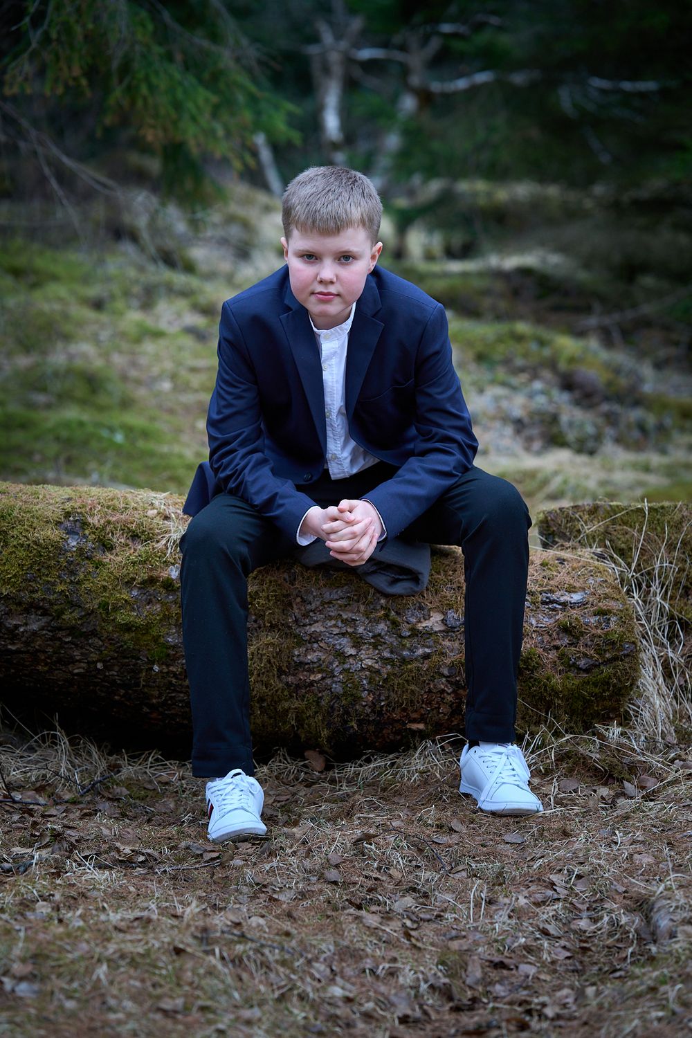 A person in a navy suit and white sneakers sits thoughtfully on a mossy log in a forest setting.