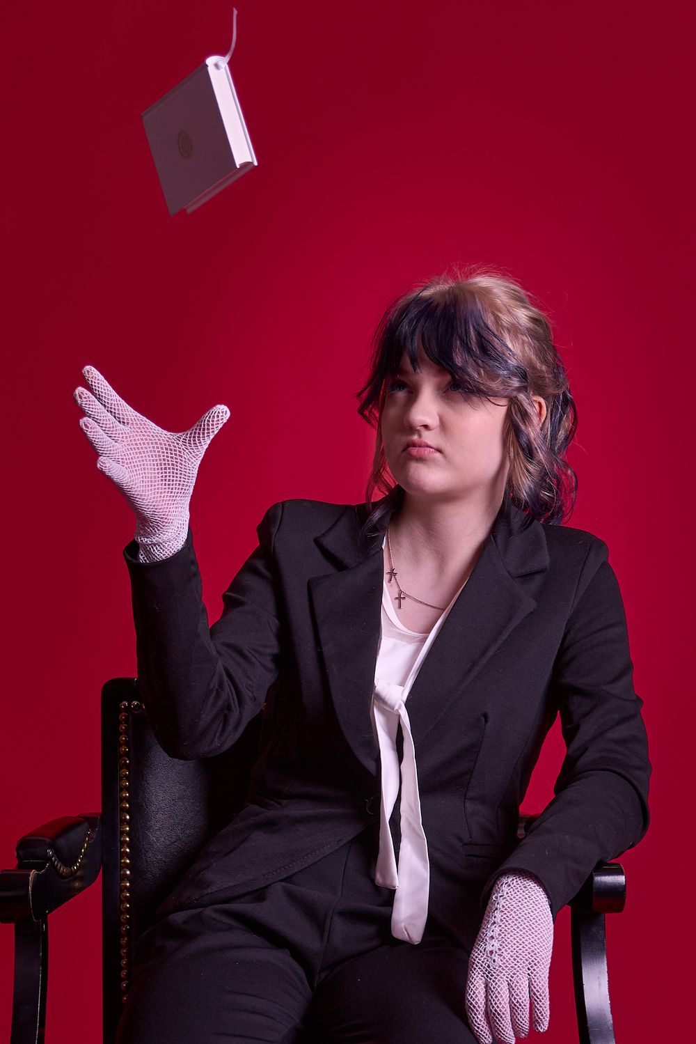 A person in a black suit and white gloves tosses a small object while seated in an elegant armchair against a red backdrop.