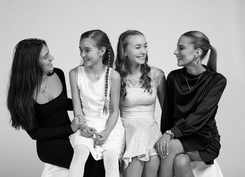 Four young friends laughing together while sitting in a row wearing casual black and white outfits in a studio setting.