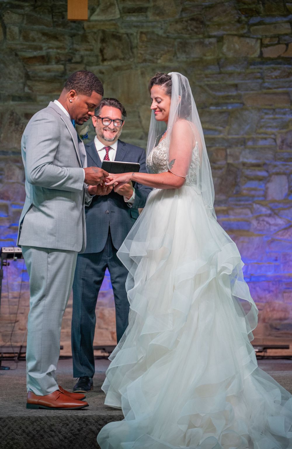 A wedding ceremony moment showing the exchange of rings in a stone-walled venue with blue uplighting.