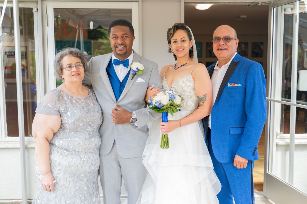 A group poses for a formal wedding photo outside glass doors, with two wearing gray and two in blue attire.
