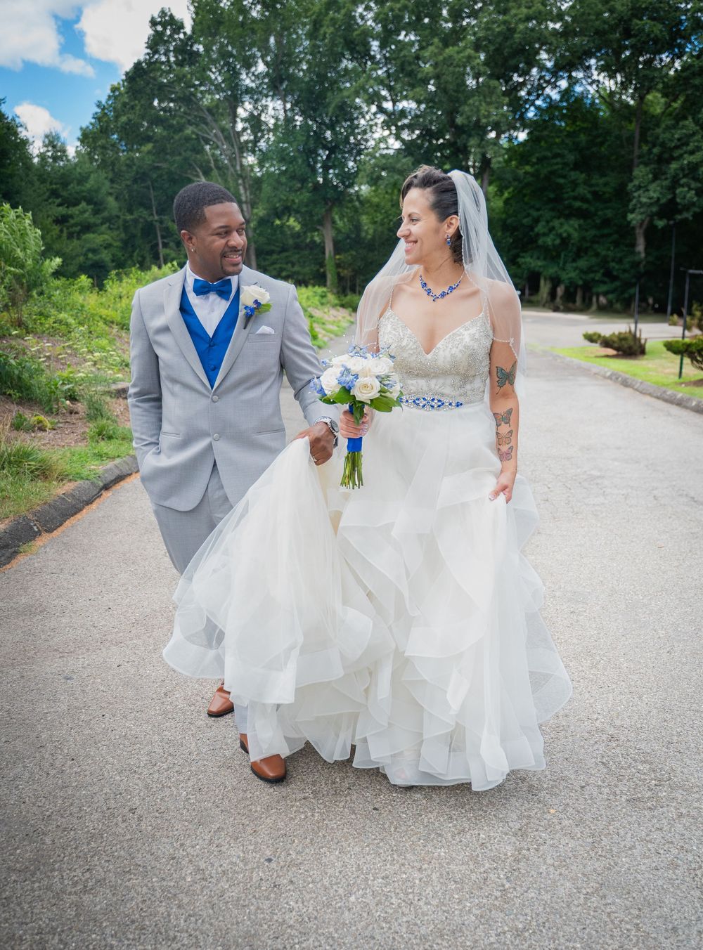 A couple in wedding attire walks hand in hand along a winding gravel path surrounded by lush greenery.
