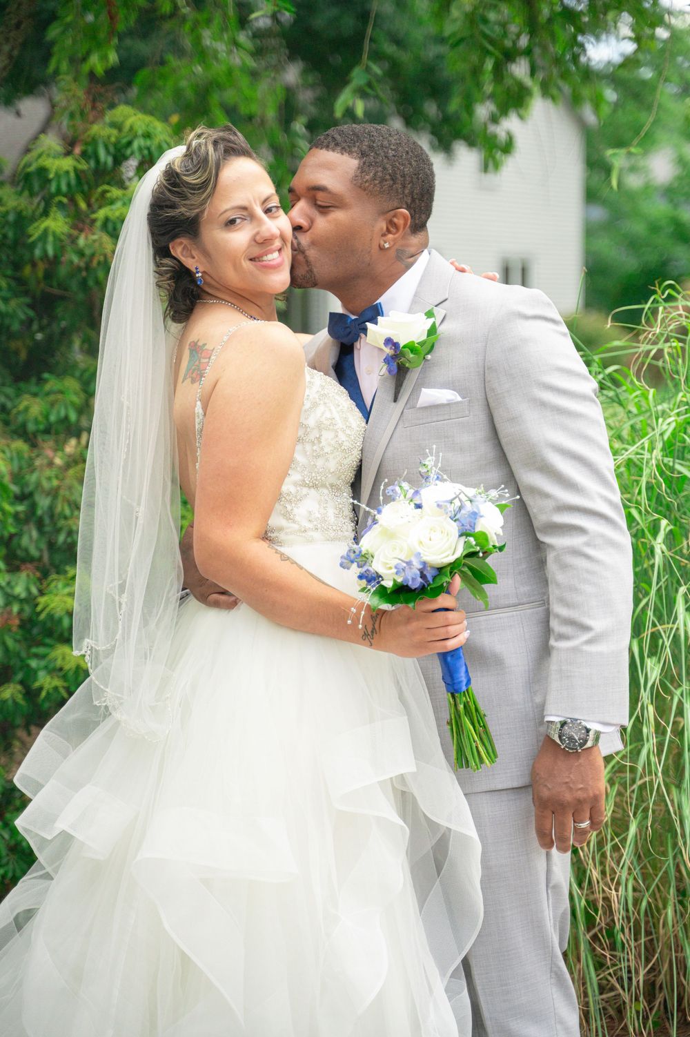 Bride and groom share intimate wedding day kisses in a lush green garden setting, wearing white dress and grey suit.