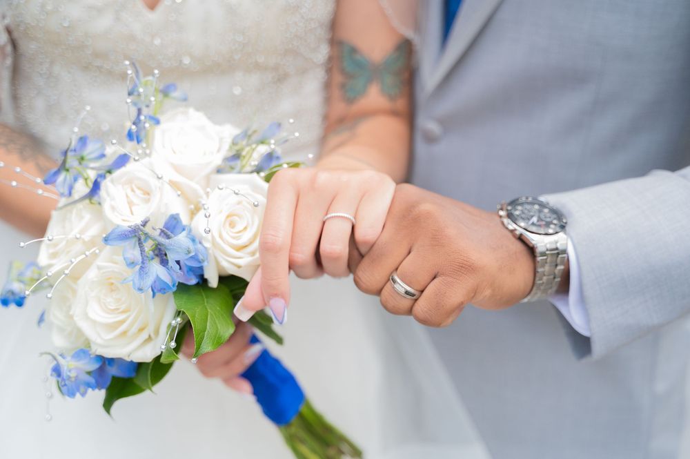 Close-up of newlyweds' hands with wedding rings next to a white and blue floral bouquet.