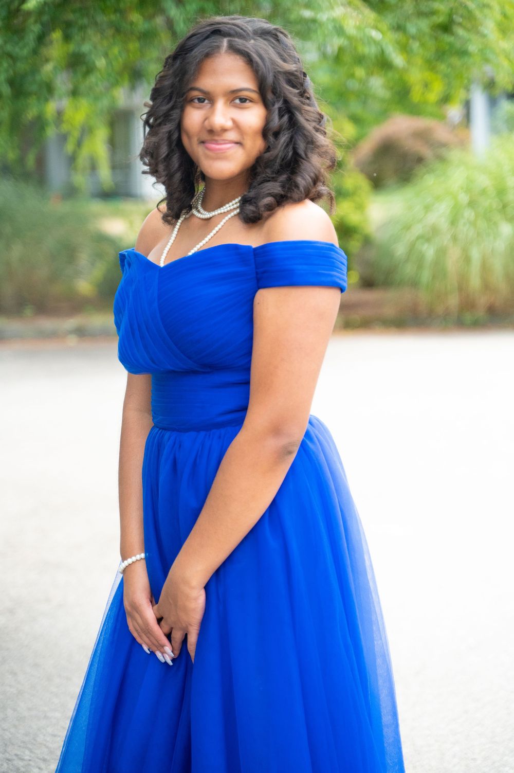 A person poses outdoors in a vibrant royal blue off-shoulder formal dress with tulle skirt against a blurred natural background.