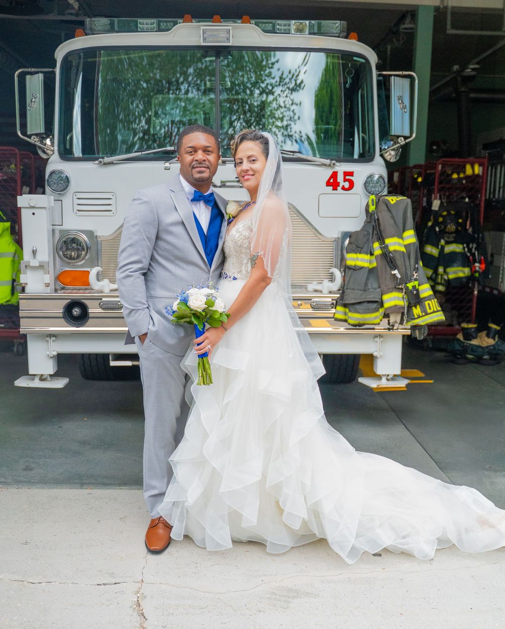 A couple poses together outside a garage with white buses, the bride wearing a flowing white gown and the groom in gray.