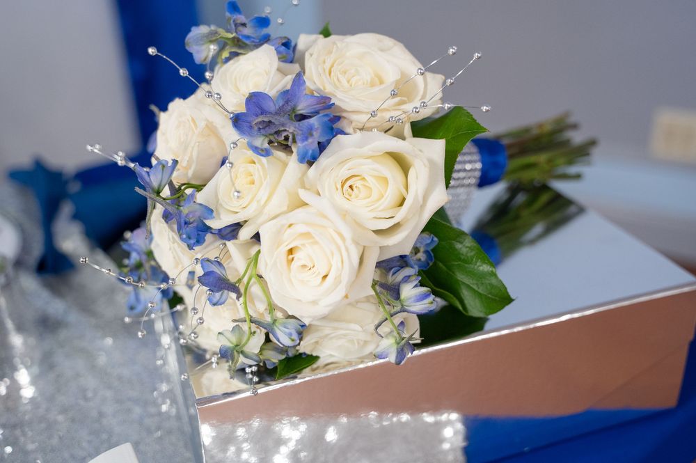 White roses and blue delphinium flowers arranged in an elegant bridal bouquet with textured stems and greenery.