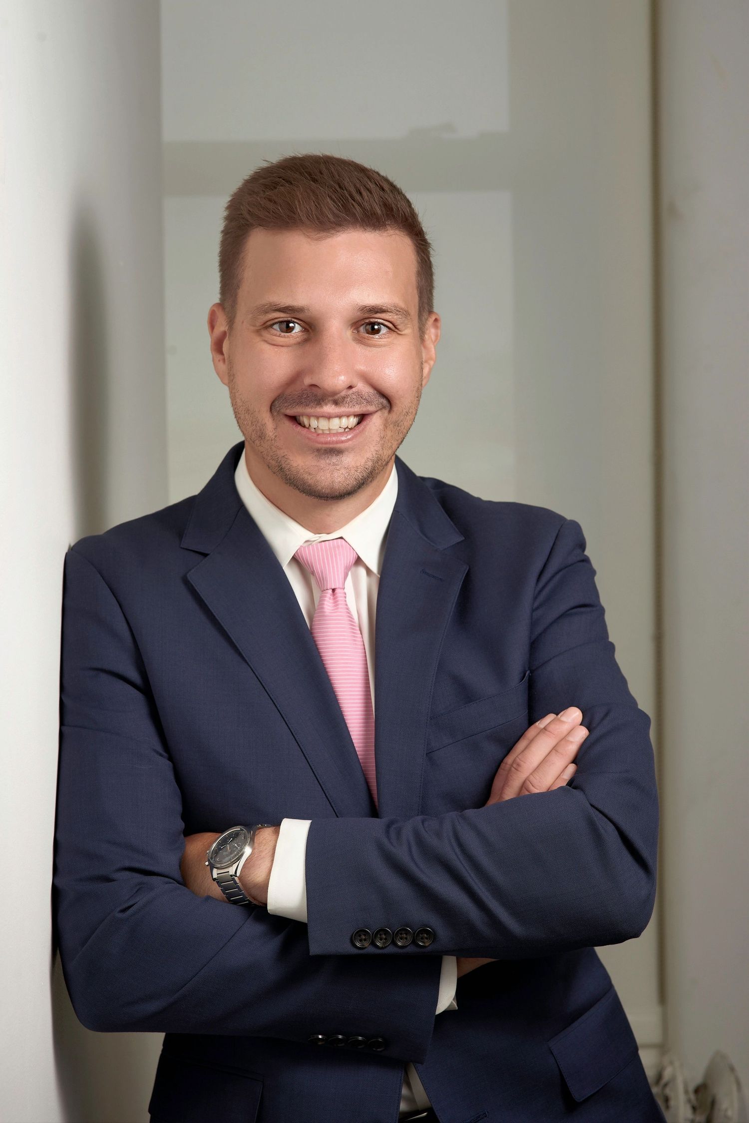 Young professional man in a navy suit and pink tie smiling with arms crossed in an office.