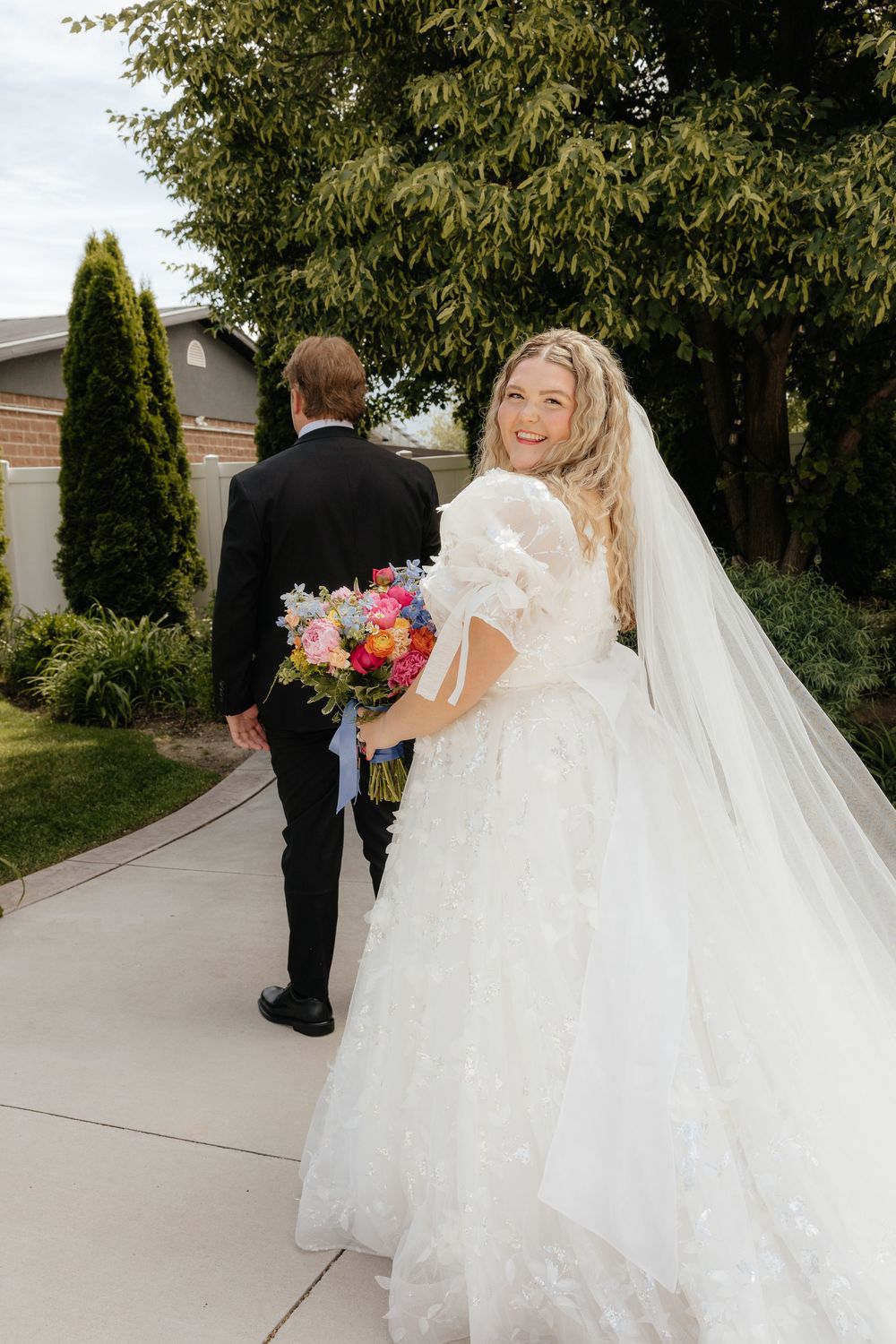 Smiling bride in white floral gown holding colorful bouquet walking with groom in black suit outdoors.