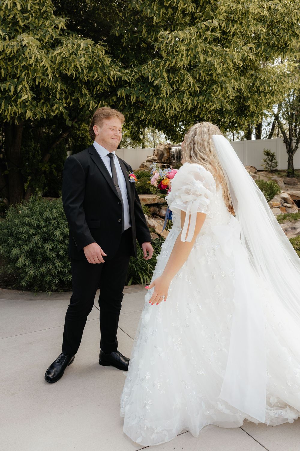 Groom in black suit smiling as bride in white floral ballgown approaches during outdoor first look.