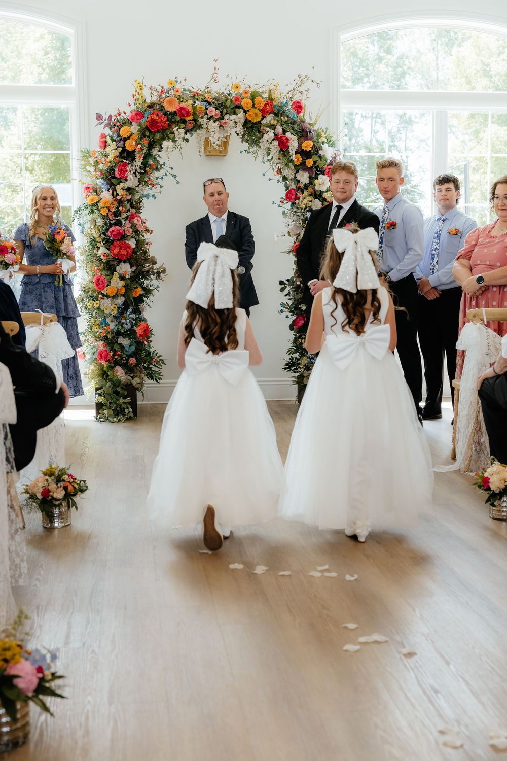 Two flower girls in white dresses with large bows walking down aisle toward floral ceremony arch.