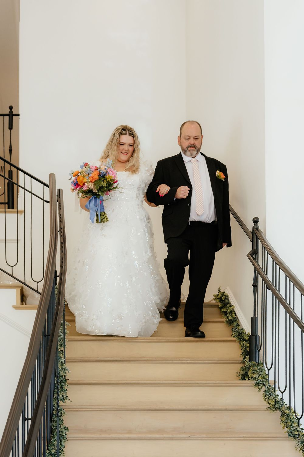 Bride in white gown holding colorful bouquet walks down staircase with father on wedding day.