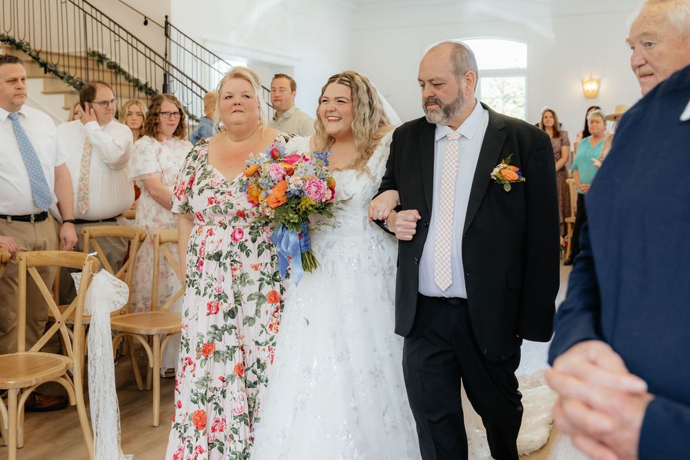 Bride escorted down aisle by parents in bright floral wedding ceremony with guests standing.