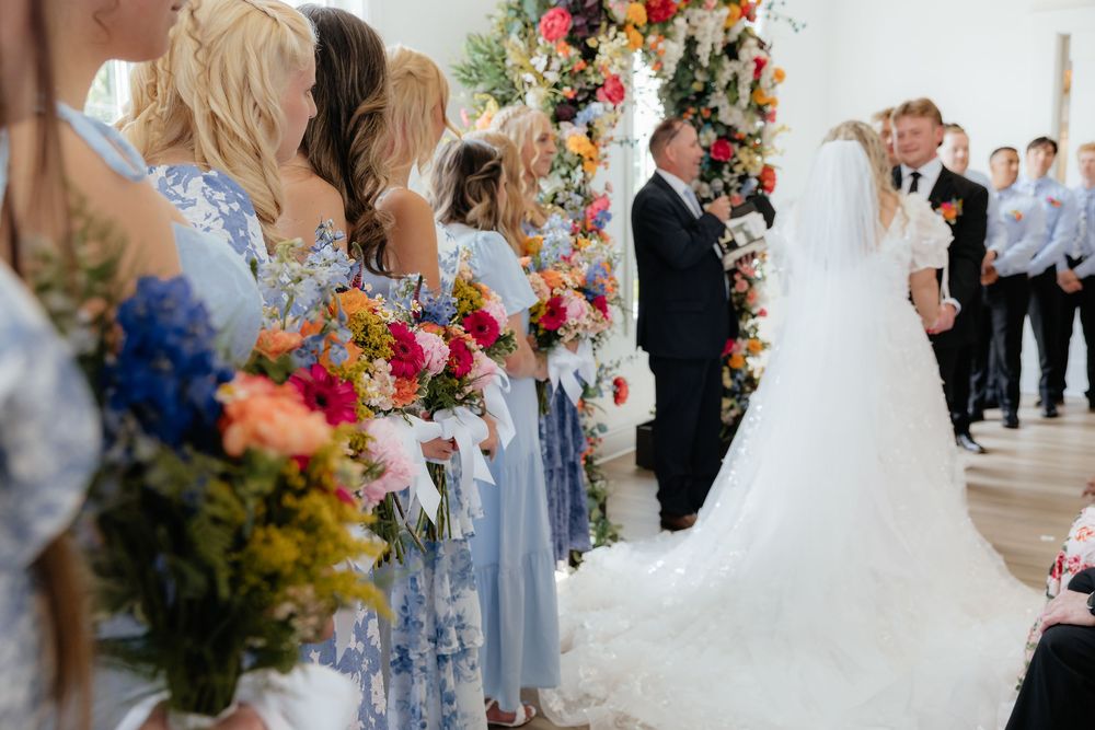 Bridesmaids in blue dresses holding colorful bouquets stand beside bride at floral arch wedding ceremony.