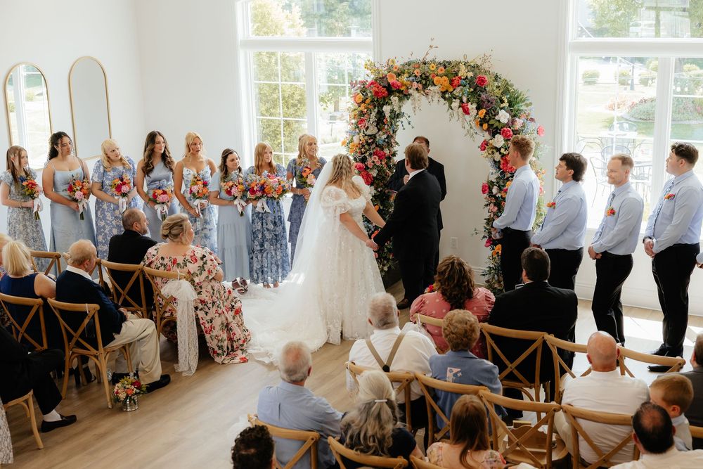 Wedding ceremony with bride and groom at floral arch altar flanked by bridesmaids and groomsmen.