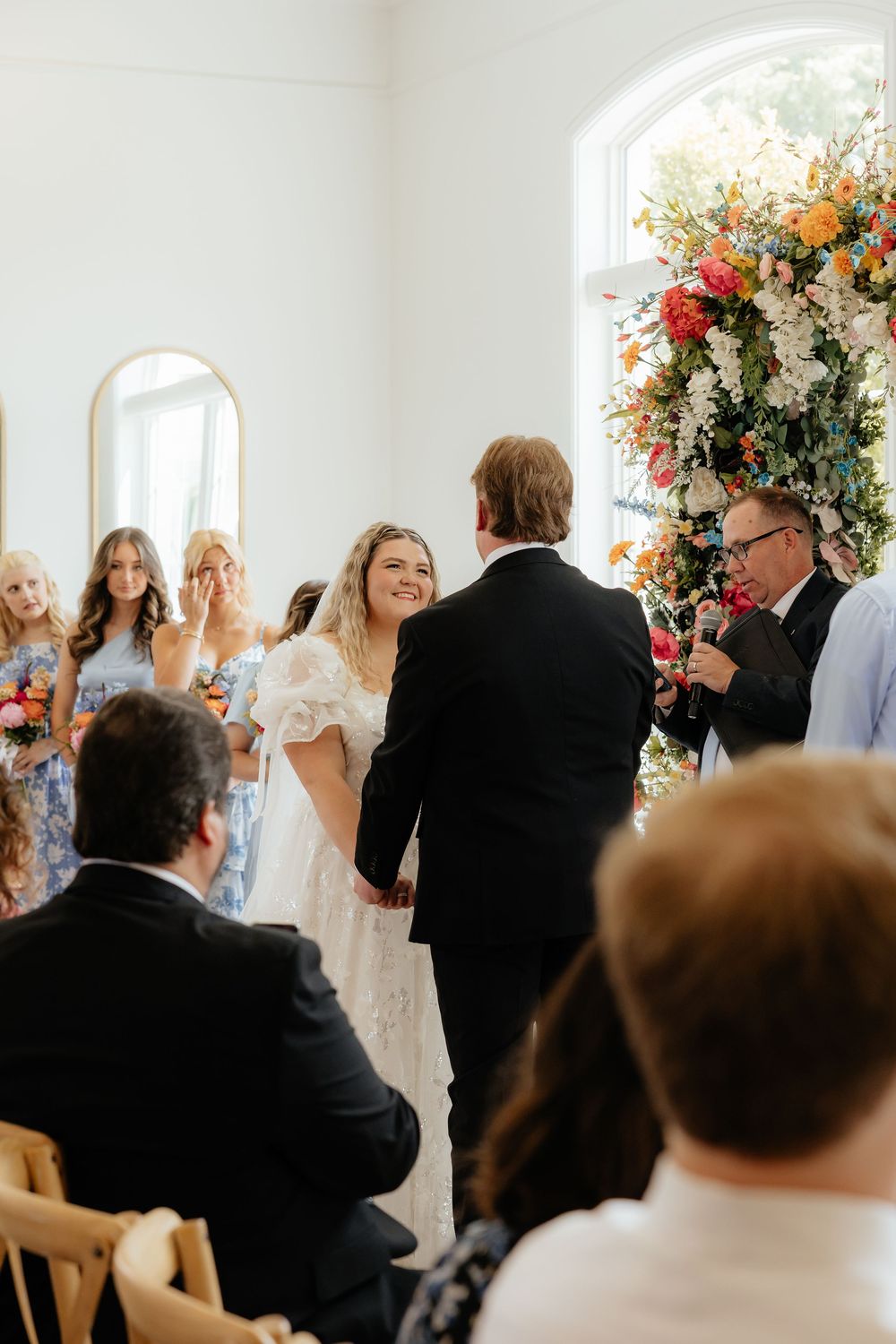 Bride and groom hold hands at altar during wedding ceremony beside vibrant floral arrangement.