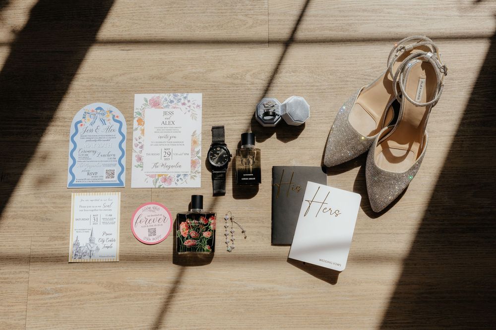 Flat lay of wedding details including floral invitations, perfume bottles, watch, and gold heels.