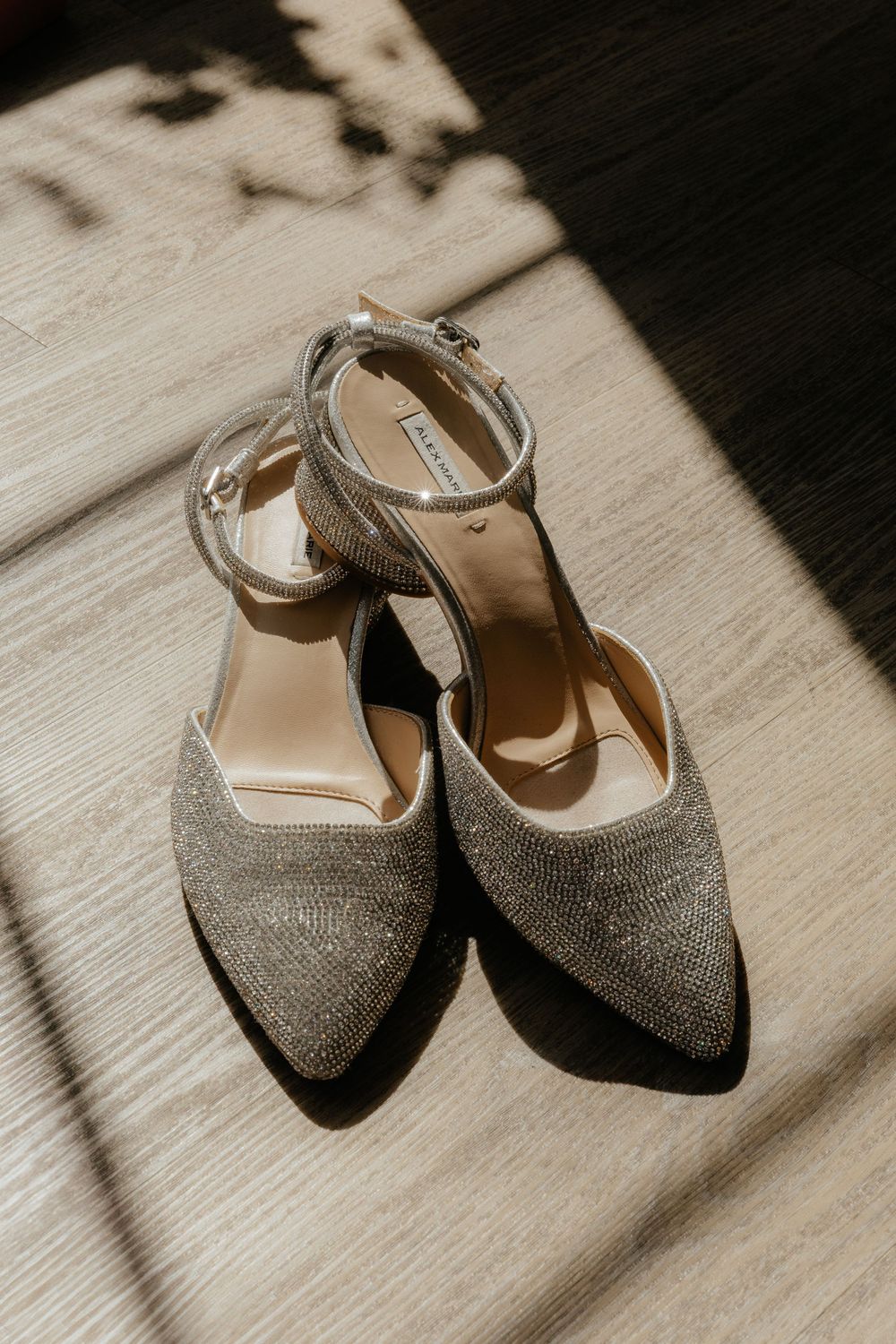Sparkling silver crystal embellished bridal heels with ankle straps on a wooden floor in sunlight.