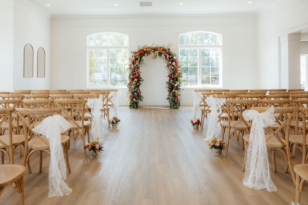 Bright wedding ceremony venue with floral arch, wooden cross-back chairs, and white ribbon decor.