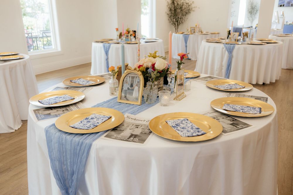 Round reception table set with gold chargers, blue runner, floral centerpiece, and vintage photos.