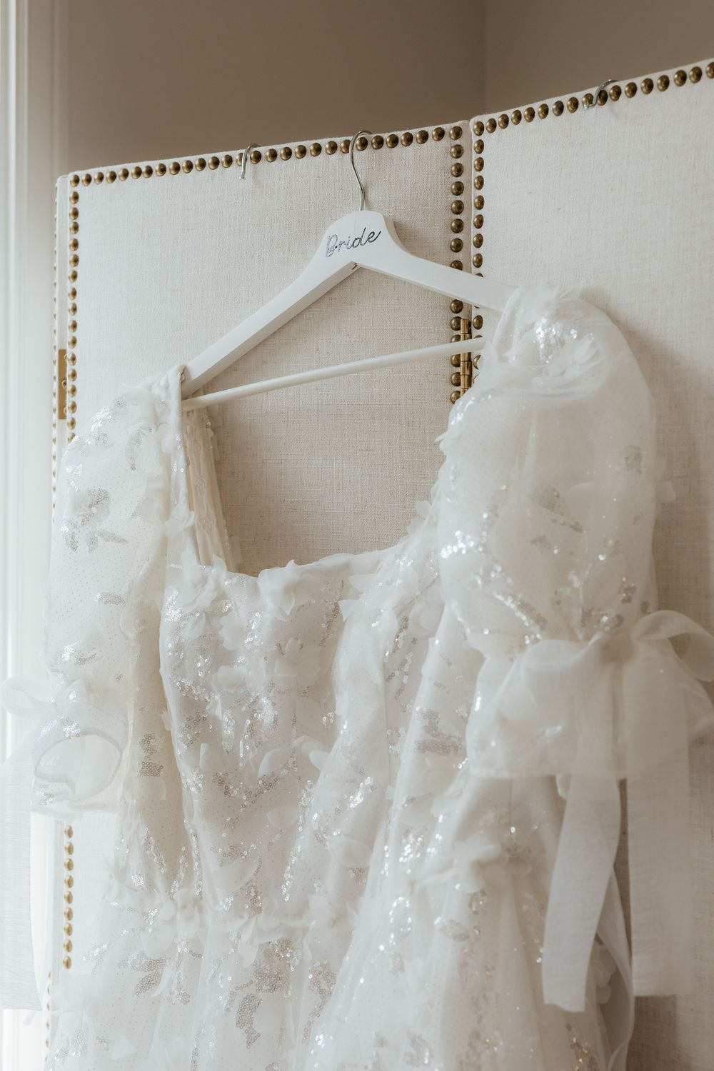 Elegant beaded white wedding dress with puff sleeves hanging on a bride hanger against a tufted headboard.