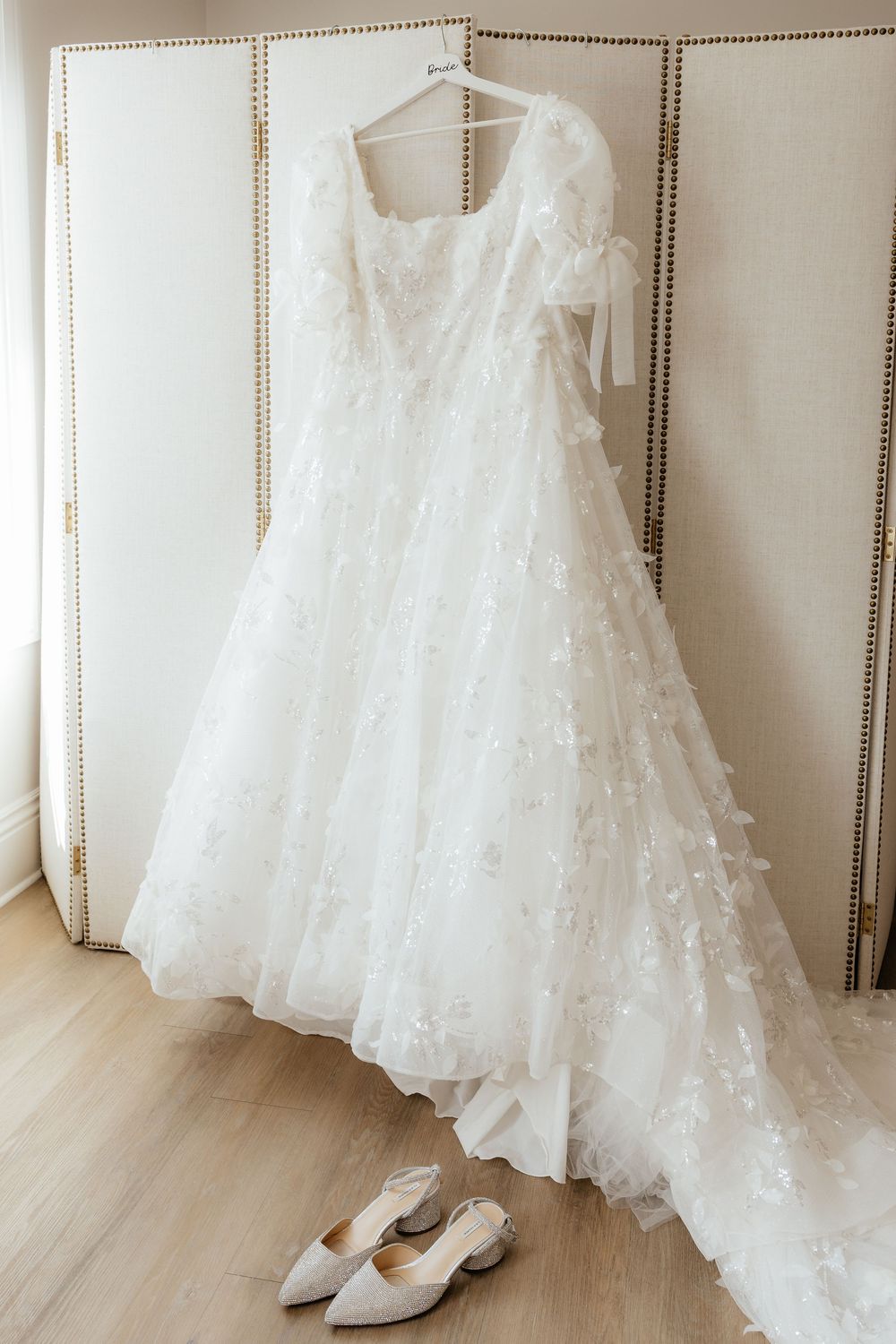 Elegant white wedding gown with 3D floral appliques hanging on a hanger with bridal heels below.